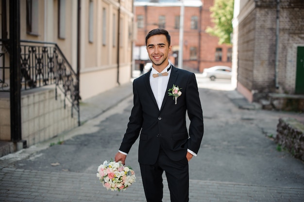 Novio en un traje negro caminando por la calle con un ramo de rosas