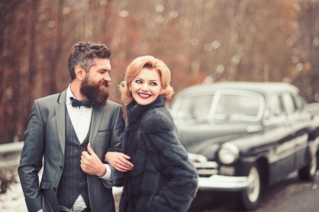 El novio con un traje negro con una mujer al aire libre cerca de un auto retro