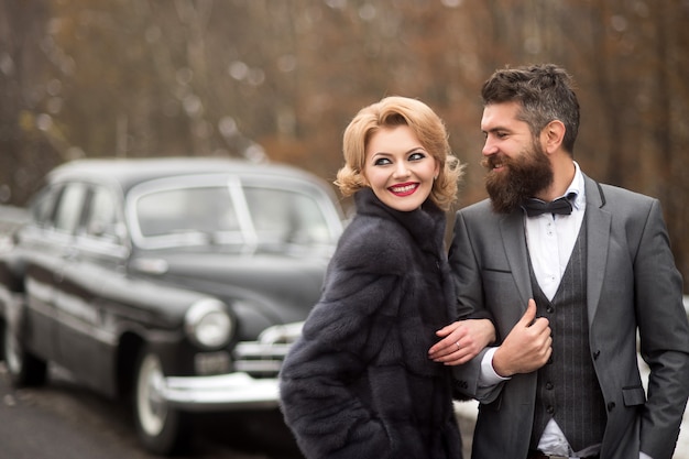 Novio con traje negro con mujer al aire libre cerca de coche retro. Cita romántica. Gente vintage