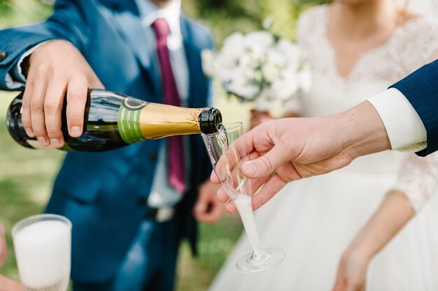 El novio vierte champán en una copa al fondo de la novia en la naturaleza