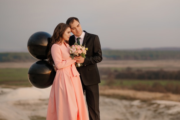 Los novios felices caminan en el hermoso paisaje al atardecer con grandes globos de helio. Concepto de boda