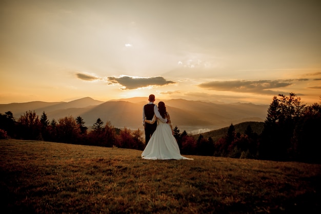 Los novios miran la puesta de sol de pie en la colina