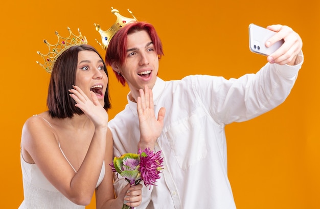 Novios novios y novios con ramo de flores en vestido de novia con coronas de oro sonriendo alegremente haciendo selfie con smartphone saludando con las manos