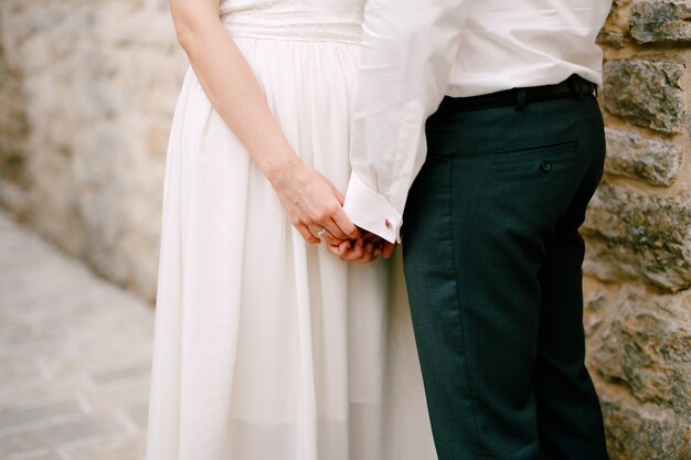 Los novios se paran cerca de un muro de piedra y se abrazan conmovedoramente las manos de los demás