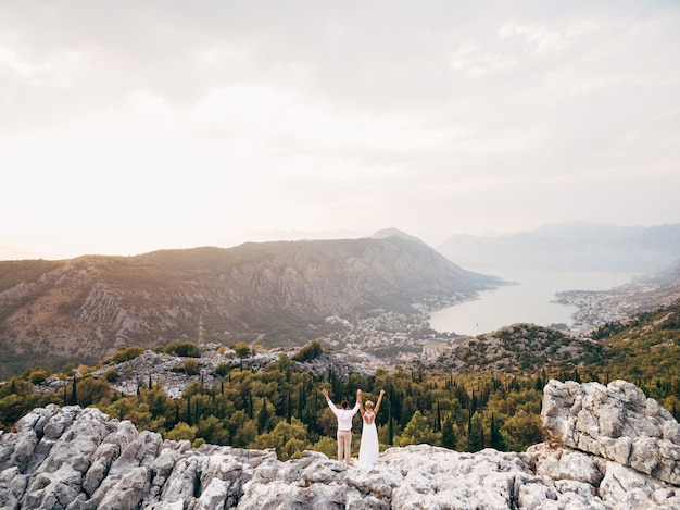 Los novios se paran en el monte lovcen con las manos en alto y miran la bahía de kotor