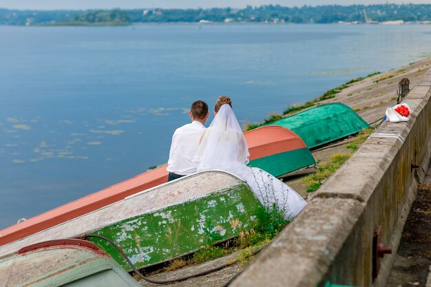 Novios sentados detrás, entre viejos barcos cerca del río.