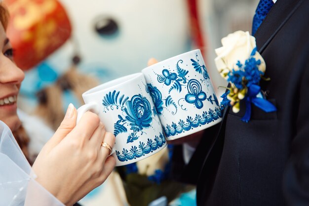 Foto novios sosteniendo tazas de té con motivos azules del folklore ruso. boda ruusiana con doble felicidad