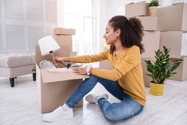 Novo apartamento. menina agradável de cabelos encaracolados abrindo a caixa de um abajur e sorrindo, tendo se mudado para um novo apartamento