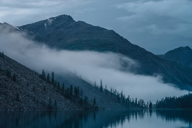 Nube baja sobre el lago de las tierras altas. Siluetas de árboles en la ladera a lo largo del lago de montaña en una densa niebla. Reflejo de pinos a aguas tranquilas. Tranquilo paisaje alpino temprano en la mañana. Paisaje atmosférico fantasmal