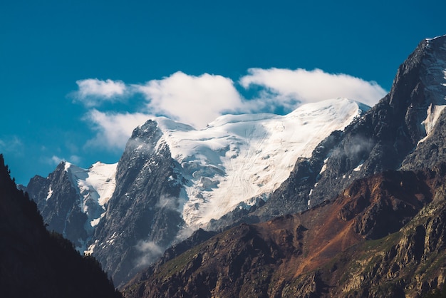 Nubes bajas y niebla en la cima de la cordillera. Glaciar bajo el cielo azul claro. Pico de montaña cubierto de nieve en un día soleado. Canto rocoso gigante con nieve. Atmosférico paisaje de montaña minimalista de naturaleza majestuosa.