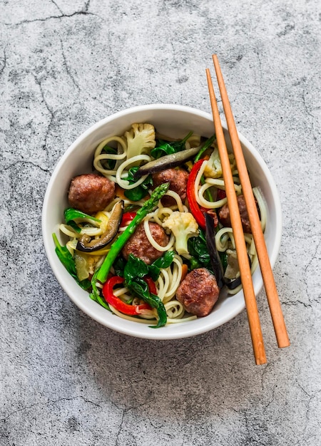 Nudeln mit Fleischbällchen und Gemüse Blumenkohl-Zucchini-Pfeffer-Spinat-Auberginen-Karotten gebratener Wok auf einer Draufsicht des grauen Hintergrundes Beschneidungspfad eingeschlossen