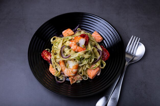 Nudeln mit Meeresfrüchten, sonnengetrockneten Tomaten, Kapern und roten Zwiebeln. Hausgemachte Pasta mit Garnelen, Lachs (Forelle) und Pesto-Sauce. Schwarzer Hintergrund, schwarze Platte. Nahansicht.