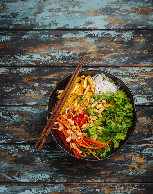 Nudelsalat im asiatischen, chinesischen oder vietnamesischen Stil mit frischem Gemüse, gebratenem Tofu und Erdnüssen, serviert in rustikaler Keramikschale auf buntem Holzhintergrund. Gesunde Ernährung, sauberes Essen oder veganes Konzept