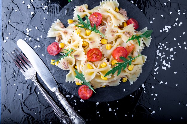 Nudelsalat in der Schieferplatte mit Tomaten Kirsche, Thunfisch, Mais und Rucola in der Nähe von Messer und Löffel. Ansicht von oben. Zutaten. Italienisches Essen.