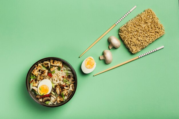 Nudelschale mit Pilzen, Ei, Tofukäse, Stäbchen und Zutaten auf grünem Grund. Asiatisches Lebensmittelkonzept.