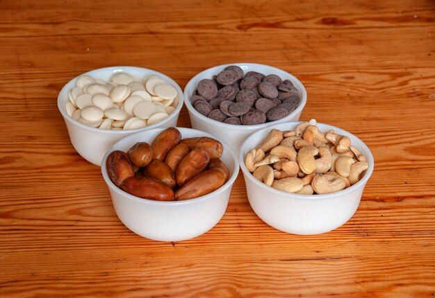 Nueces de anacardo, dátiles, gotas de chocolate blanco y con leche en tazones pequeños blancos.