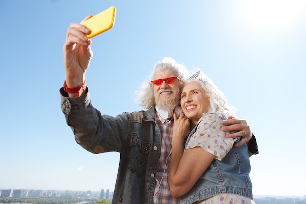 Nuestra foto. Hombre positivo alegre sosteniendo un teléfono inteligente mientras toma una foto de él y su esposa