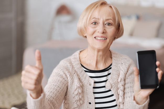 Nützlich und unterhaltsam. Emotional aufgeregte reife Frau, die auf einem Sofa sitzt und ihr neues Smartphone zeigt, während sie ihren Daumen hochhält