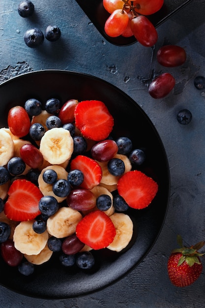 Nützliches Frühstück mit Früchten und Beeren Draufsicht in Scheiben geschnittene Bananen Erdbeeren Trauben und Blaubeeren in einer schwarzen Platte