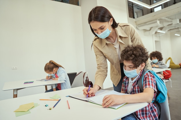 Nueva maestra moderna normal con mascarilla protectora ayudando a un niño en el aula de niños