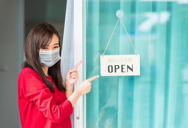 Nueva mujer joven asiática normal que usa mascarilla protege el dedo alegre para notar la etiqueta de tablero de madera "BIENVENIDA ABIERTA" colgada a través de la puerta de vidrio de la tienda principal, el negocio se abre después de la pandemia del coronavirus