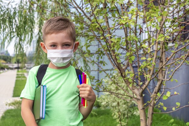 Nueva normalidad, regreso a clases. El colegial con máscara médica y mochila sosteniendo libros de texto y rotuladores al aire libre