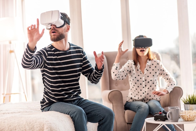 Nueva realidad. Hombre agradable bastante emocional que estira la mano mientras intenta auriculares VR y se sienta en la cama