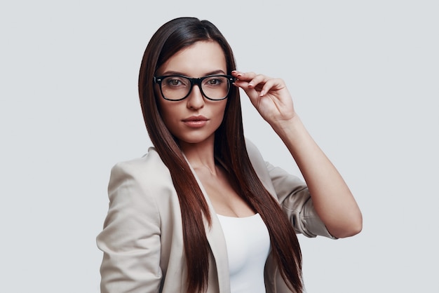 Nueva solución todos los días. Atractiva mujer joven mirando a la cámara y ajustando las gafas mientras está de pie contra el fondo gris