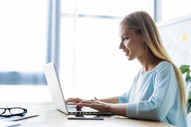 Nueva solución todos los días Mujer joven segura con ropa informal elegante trabajando en una computadora portátil mientras está sentada en su lugar de trabajo en la oficina
