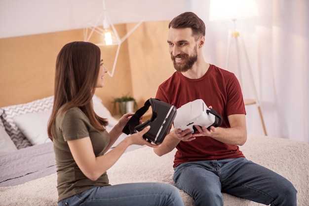 Nueva tecnología. Atractiva pareja enérgica positiva sentada en el mal mientras se miran y sostienen gafas VR