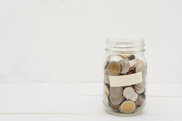 Nuevas monedas baht tailandesas en un frasco de vidrio con etiqueta en blanco sobre una mesa de madera blanca con espacio para copiar