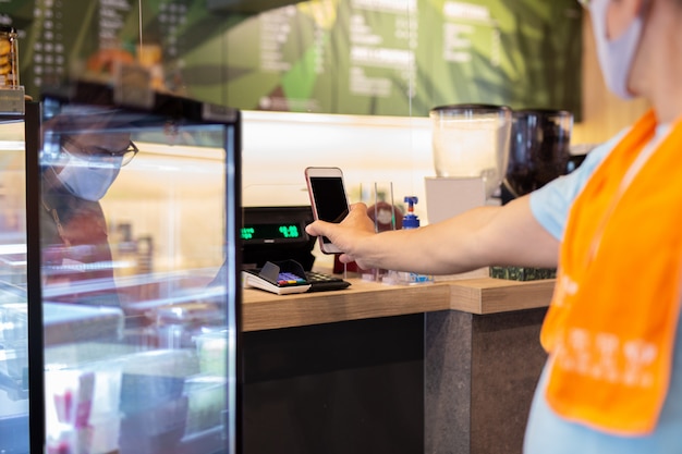 Nuevo cliente masculino nomarl que paga la factura por teléfono celular con el personal antes de la partición de plástico en el café.