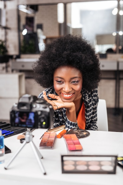 Nuevo estilo. Mujer sonriente con un top naranja demostrando maquillaje nuevo mientras realiza un tutorial en línea