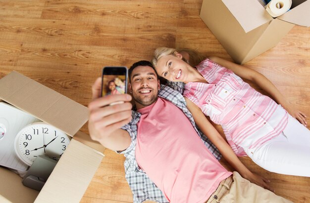 nuevo hogar, tecnología, gente, reparación y concepto de mudanza - pareja feliz tomándose selfie con un smartphone y tirados en el suelo entre cajas de cartón en casa