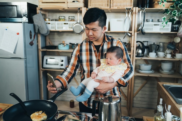 el nuevo padre asiático japonés que sostiene un teléfono inteligente está tomando una foto con su linda niña en el brazo mientras cocina el desayuno en la cocina en casa