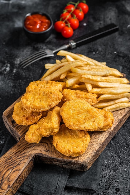 Nuggets de pollo con salsa de tomate y papas fritas sobre plancha de madera. Fondo negro. Vista superior.