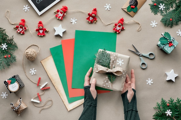 Null Abfall Weihnachten, flache Lage, Draufsicht auf Bastelpapierwand mit roter Puppengirlande, weibliche Hände, die Geschenk auf Farbpapierstapel einwickeln. Umweltfreundliche handgemachte Weihnachtsdekorationen ohne Kunststoff