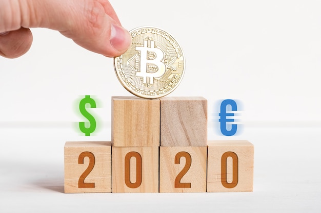 Números em cubos de madeira em um fundo branco ao lado de uma moeda de bitcoin e sinais de dólar e euro.