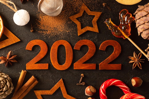 Números de madera marrón 2022 sobre un fondo oscuro. Hay estrellas talladas en madera, especias, cacao esparcido y una taza con malvaviscos alrededor.