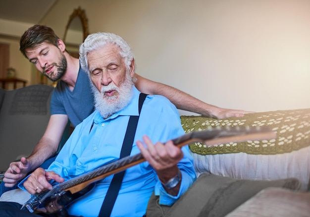 Nunca se es demasiado viejo para convertirse en una estrella de rock Foto de un joven feliz enseñando a su anciano abuelo a tocar la guitarra eléctrica en el sofá de su casa