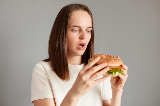 Foto nutrição adequada comida rápida saudável escolha insalubre jovem mulher atônita espantada vestindo camiseta branca segurando hambúrguer na mão isolado em fundo cinza