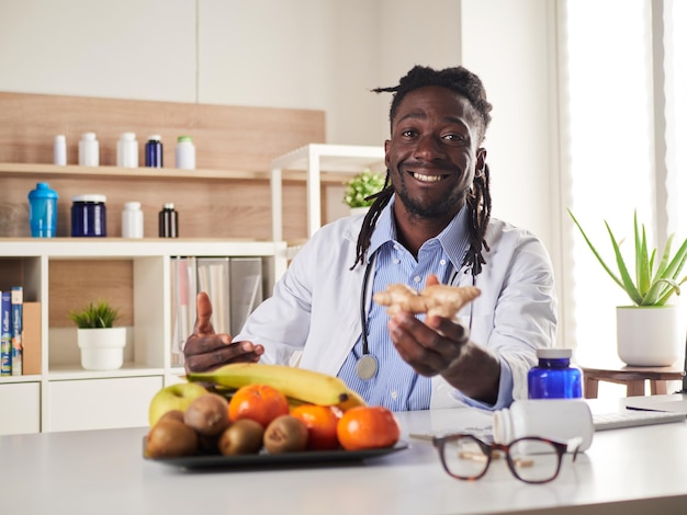 Nutricionista afroamericana mirando a la cámara y mostrando jengibre saludable en la consulta