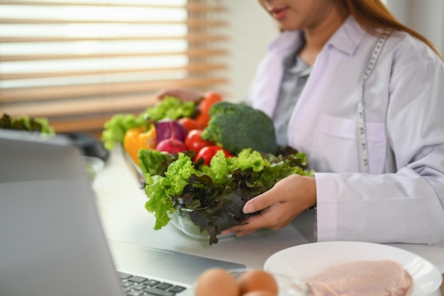 Nutricionista con una bandeja de vidrio de verduras frescas promoviendo una dieta saludable durante la consulta en línea