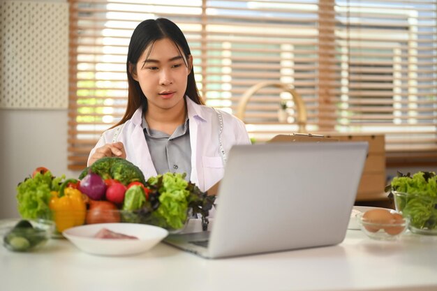 Nutricionista en bata blanca llamando al paciente y dando consultas en línea en una computadora portátil
