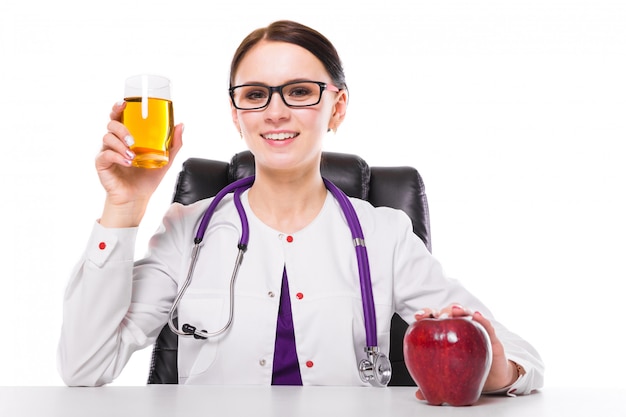 Nutricionista femenina sentada en su lugar de trabajo mostrando y ofreciendo un vaso de jugo fresco de manzana con manzana en la mano en blanco