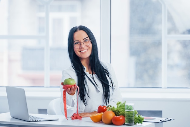 Nutricionista feminina de jaleco branco segurando uma maçã com fita métrica.