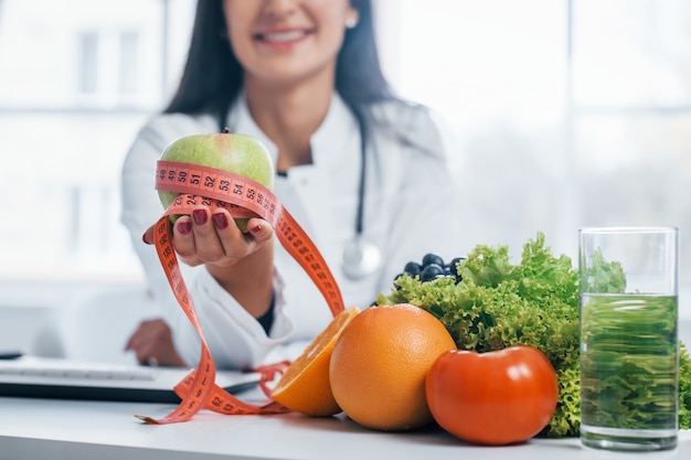 Foto nutricionista feminina de jaleco branco, sentado dentro de casa, no escritório no local de trabalho.