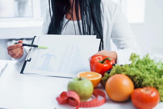 Nutricionista mujer en bata blanca sentada en el interior de la oficina en el lugar de trabajo y mostrando información sobre el documento.