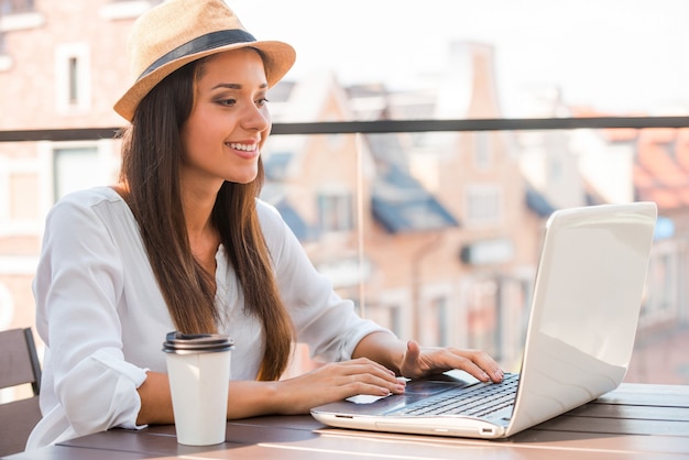 Nutzen Sie die Vorteile von kostenlosem WLAN. Schöne junge Frau mit flippigem Hut, die am Laptop arbeitet und lächelt, während sie draußen sitzt