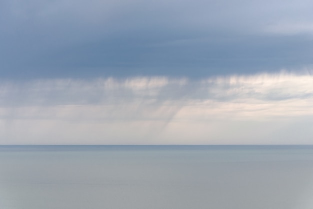 Nuvens chuvosas sobre um mar calmo, faixa de chuva no horizonte, foco suave, longa exposição, paisagem abstrata.